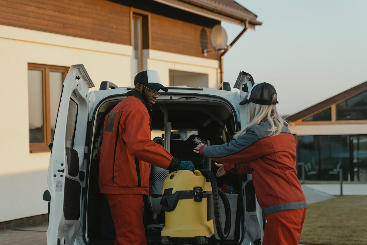 about-us Two cleaners in red uniforms load a vacuum into a white van outdoors.